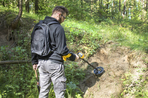 Homem com uma mochila nas costas fica na floresta com um detector de metais na mão e procura na área em busca de tesouros
. - Foto, Imagem