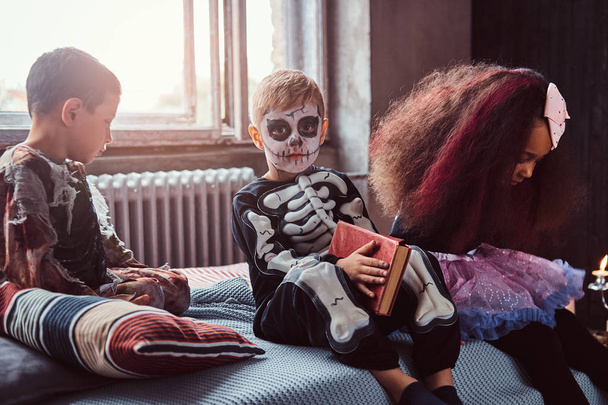 Három többnemzetiségű gyerekek olvasás rémtörténetek ülve, ágyban, egy régi ház ijesztő jelmezek. - Fotó, kép