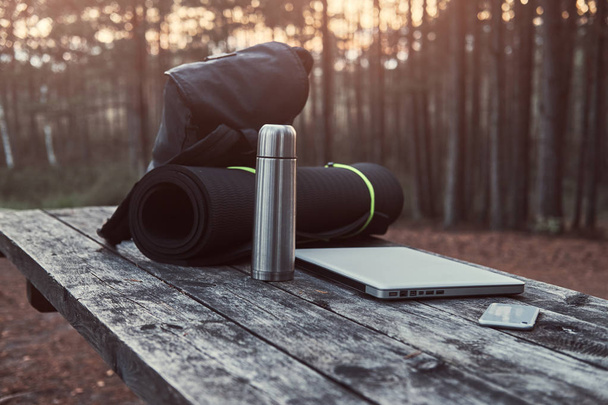 Concetto di viaggio. Zaino, tappetino turistico, laptop, smartphone e thermos bottiglia su un tavolo di legno in una bellissima foresta
. - Foto, immagini