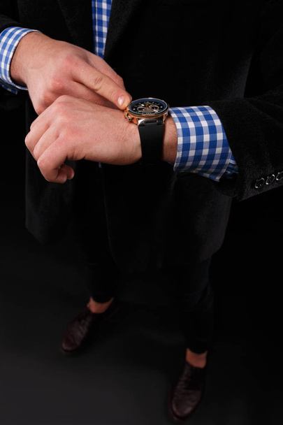 Close-up of a stylish leather black wrist watch on a male hand. A businessman in an elegant costume with a gold watch on a black background. - Фото, изображение