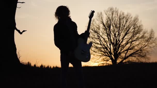 Mies soittaa sähkökitaraa ja laulaa laulun pellolla lähellä puuta auringonlaskun aikaan. siluetti
 - Materiaali, video