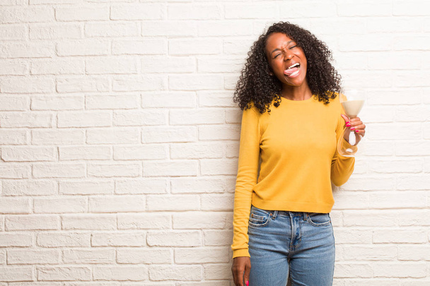 Jeune femme noire avec verre de sable expression de confiance et d'émotion montrant la langue contre le mur de briques
 - Photo, image