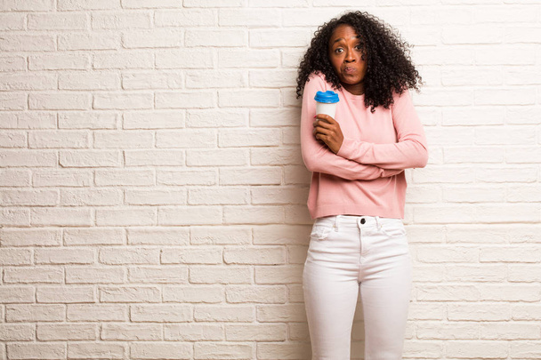 Молода чорна жінка з одноразовою чашкою кави сумнівається і плече на цегляній стіні
 - Фото, зображення