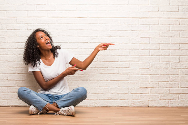 Giovane donna nera seduta su un pavimento di legno che punta a lato contro muro di mattoni
 - Foto, immagini