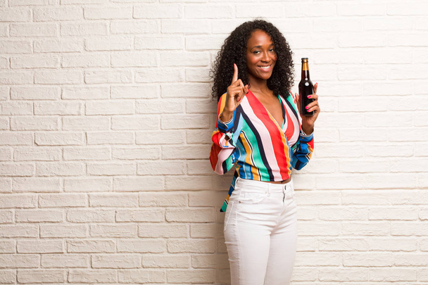 Νεαρή μαύρη γυναίκα σε πολύχρωμο πουκάμισο με μπουκάλι μπύρας που δείχνει στον αριθμό ένα τούβλο τοίχο - Φωτογραφία, εικόνα
