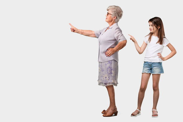Volledige lichaam van een bejaarde dame en haar kleindochter wijst naar de kant, glimlachend verrast presenteren iets, natuurlijke en ongedwongen - Foto, afbeelding