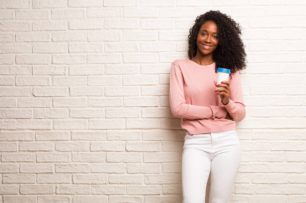 Young black woman with disposable coffee cup cheerful against brick wall - Photo, Image