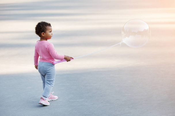 Μικρό παιδί Africanamerican σε μια ροζ πουλόβερ και γκρι παντελόνι είναι το περπάτημα στην άσφαλτο. - Φωτογραφία, εικόνα