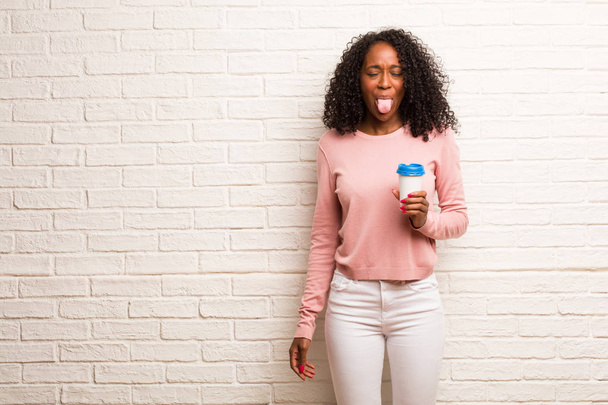 Jovem mulher negra com copo de café descartável mostrando língua contra parede de tijolo
 - Foto, Imagem
