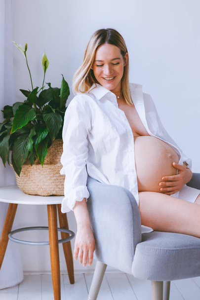 schwangere Mutter Lebensstil-Konzept, Bob Frisur blonde Frau Gesundheitsversorgung. Die Mutter sitzt auf dem grauen Sessel im Zimmer mit Scheuklappen und Holzboden. grüne Pflanze in der Ecke - Foto, Bild