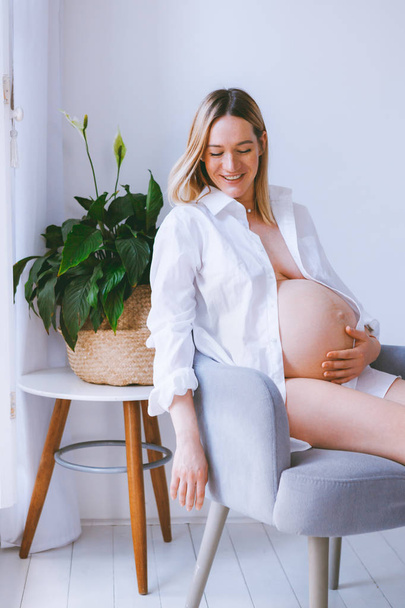 schwangere Mutter Lebensstil-Konzept, Bob Frisur blonde Frau Gesundheitsversorgung. Die Mutter sitzt auf dem grauen Sessel im Zimmer mit Scheuklappen und Holzboden. grüne Pflanze in der Ecke - Foto, Bild
