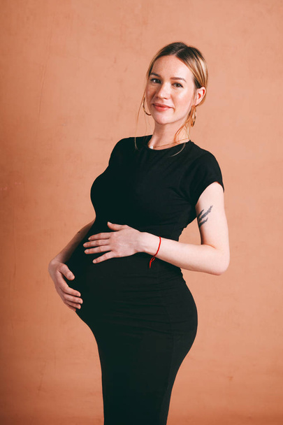classic portrait of cute beautiful pregnancy with pretty pregnant blond woman in black dress on a brown background, collected hair - Foto, Imagen