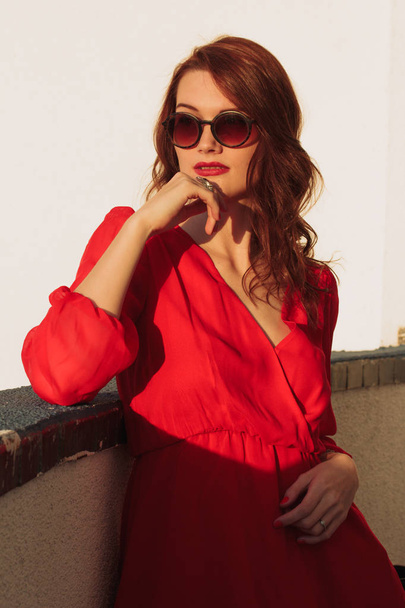 Young girl brunette standing on the balcony at sunset in a red dress and sunglasses - Фото, зображення