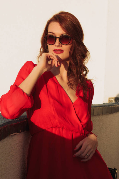 Young girl brunette standing on the balcony at sunset in a red dress and sunglasses - Φωτογραφία, εικόνα