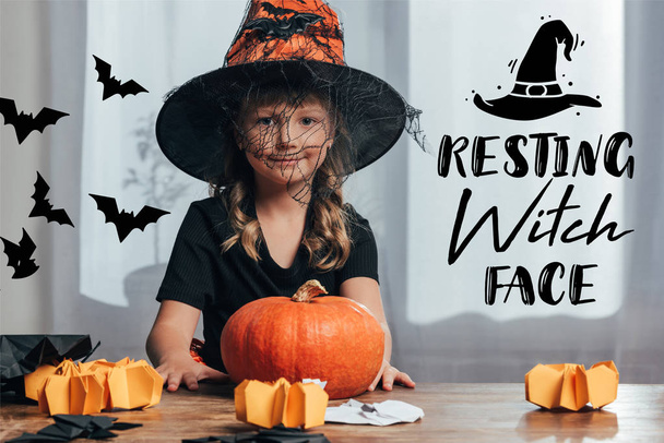 portrait of adorable kid in witch halloween costume sitting at table with pumpkin at home with "resting witch face" lettering - Photo, Image