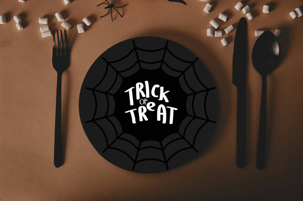 top view of black plate with spider web and "trick or treat" lettering, fork and knife with marshmallows on table, halloween concept - Fotografie, Obrázek