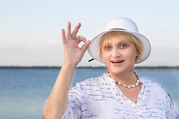 Happy smiling retired elderly lady woman in white hat near lake shows okey geste - Photo, Image