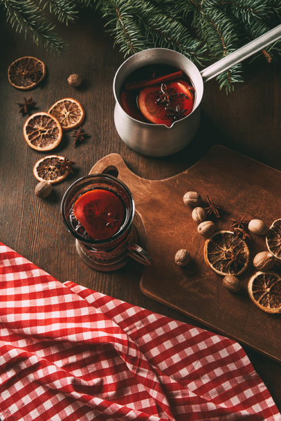homemade hot spiced wine in glass cup and saucepan with dried orange slices, nutmeg seeds and anise stars on wooden background  - Photo, Image
