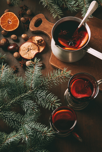 hot mulled wine in saucepan and glass cups with spices on wooden background with fir branches - Foto, imagen