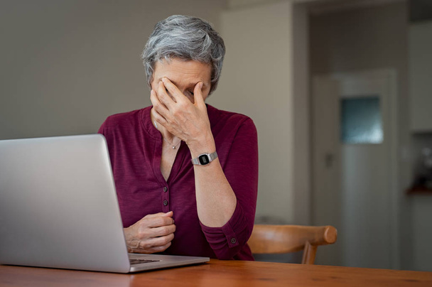 Mature businesswoman suffering a stress headache sitting at her desk with closed eyes in pain. Senior woman thinking about to complete work task. Depressed tired mature lady suffering from chronic daily headache from computer. - Foto, imagen