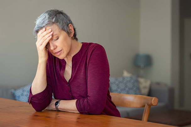 Senior woman suffering from headache while sitting at table in a living room. Depressed mature woman with head in hand thinking. Stressed old lady suffering from migraine at home. - Фото, изображение