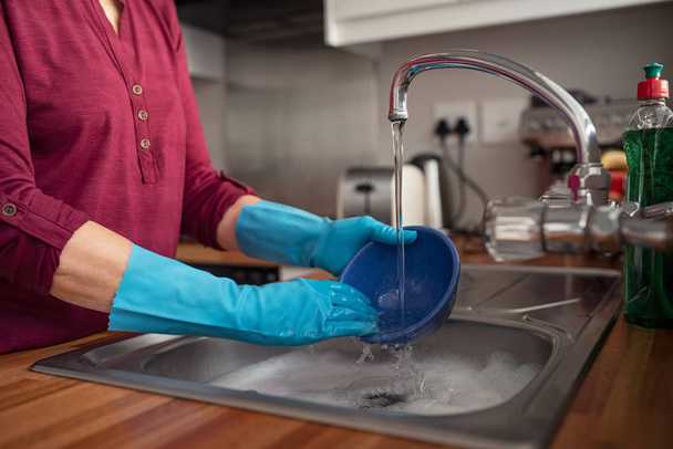 Closeup of woman hands wearing blue rubber gloves rinsing utensils using tap water in kitchen. Detail of female hands washing containers on sink. Woman in red shirt in kitchen rinsing blue bowl in washbasin.  - Photo, image