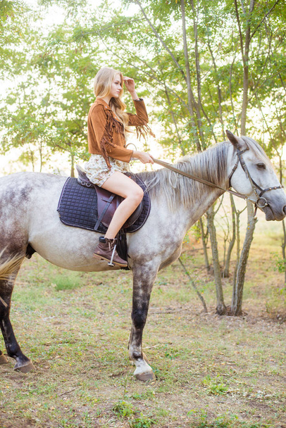 Beautiful  young girl with long blond hair in a suede jacket with a fringe smiling and astride a horse in sunset in autumn - Fotó, kép