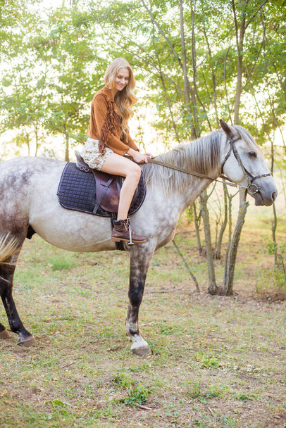 Beautiful  young girl with long blond hair in a suede jacket with a fringe smiling and astride a horse in sunset in autumn - Fotografie, Obrázek