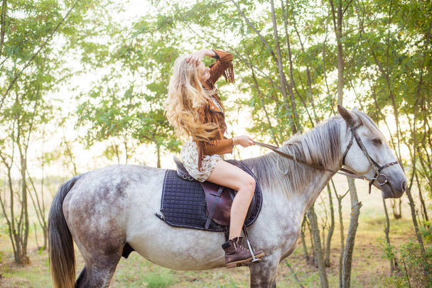 Beautiful  young girl with long blond hair in a suede jacket with a fringe smiling and astride a horse in sunset in autumn - Fotó, kép
