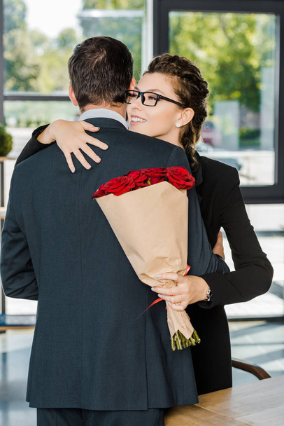 Glimlachende zakenvrouw holding boeket van rode rozen en knuffelen zakenman in kantoor - Foto, afbeelding