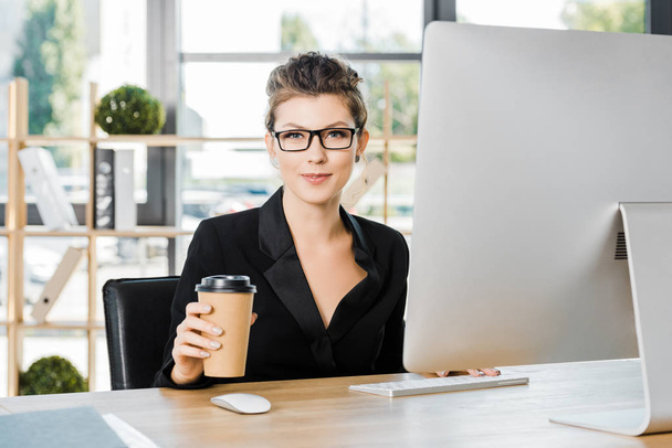 attractive businesswoman in glasses working with computer at table in office and holding disposable coffee cup - Foto, imagen