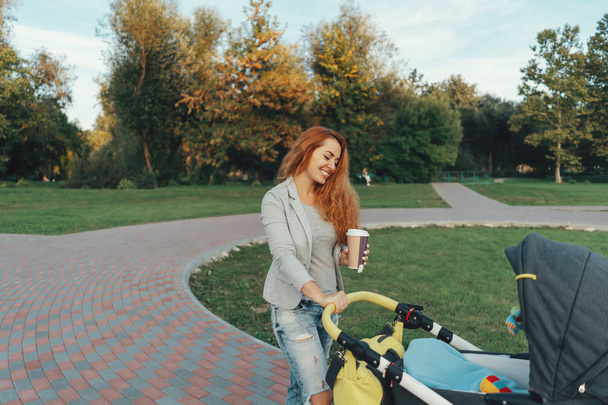 Молода мама, насолоджуючись парком, гуляє вранці, п'є гарячий чай, а її маленька дитина розслабляється на дитячому візку
. - Фото, зображення