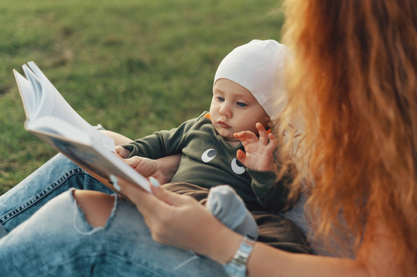 Όμορφη μητέρα είναι διαβάζοντας ένα βιβλίο με το χαριτωμένο μωρό αγόρι και χαμογελώντας, ενώ κάθονται στο γρασίδι - Φωτογραφία, εικόνα