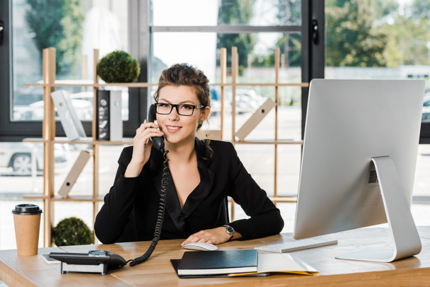 smiling attractive businesswoman talking by stationary telephone in office and looking at camera - Photo, Image