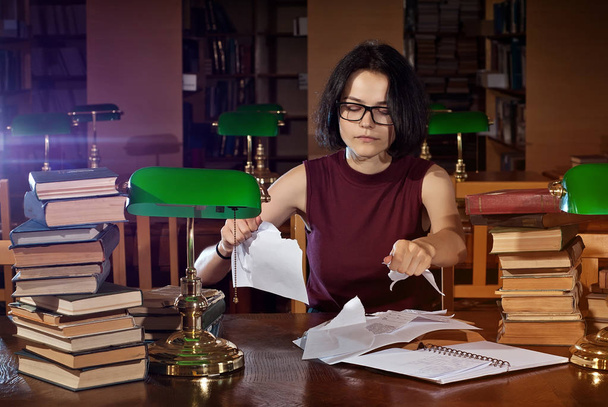 brunette in glasses at the table with books. Library with green lights on the tables. An old library with brown furniture. - Foto, imagen
