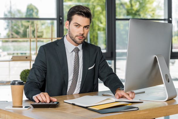 handsome businessman using calculator in office, open folder on tabletop - Photo, Image