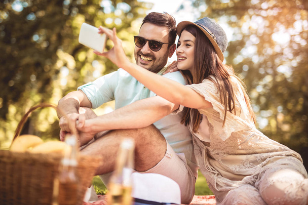 Para leży na koc piknikowy w parku z kosz piknikowy pełen owoców i zrobić selfie z telefonu - Zdjęcie, obraz