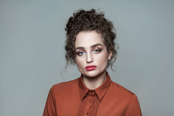 Beauty portrait of young beautiful fashion model with collected dark curly hair and nude makeup with red lips in brown shirt looking at camera serious on gray background  - Photo, Image
