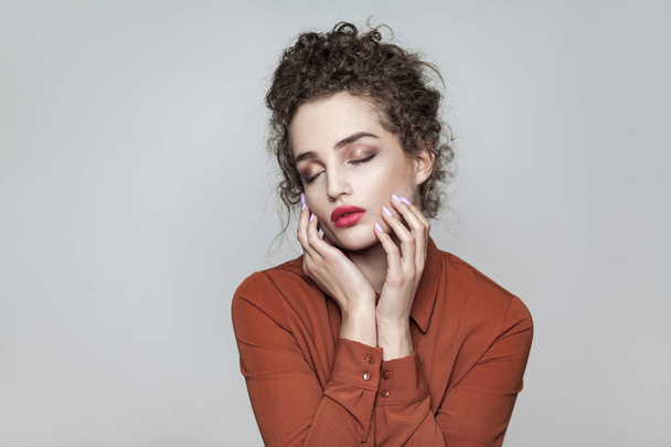 Beauty portrait of young beautiful model with collected dark curly hair and nude makeup with red lips in brown shirt touching face with closed eyes on gray background  - Foto, Imagen