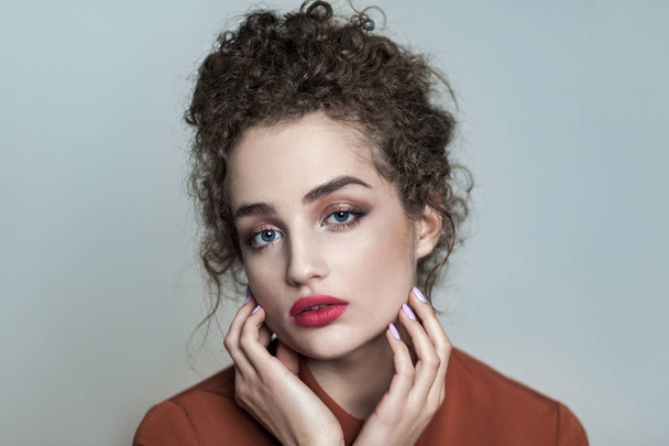 Beauty portrait of young beautiful model with collected dark curly hair and nude makeup with red lips in brown shirt looking at camera  and touching face on gray background  - Фото, зображення