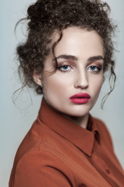 Beauty portrait of young beautiful fashion model with collected dark curly hair and nude makeup with red lips in brown shirt looking at camera serious on gray background - Foto, afbeelding