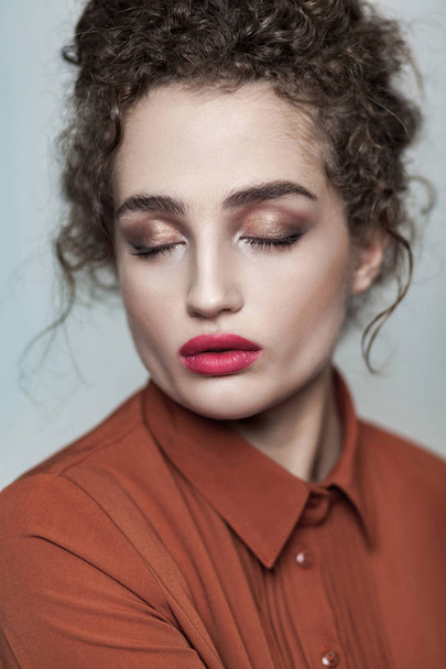 Beauty portrait of young beautiful fashion calm relaxed model with collected dark curly hair and nude makeup with red lips in brown shirt with closed eyes on gray background - Photo, Image