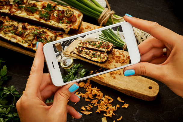woman's hands are holding the phone and taking a picture of a delicious courgette casserole on a woody table - Photo, Image