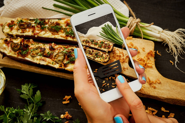 woman's hands are holding the phone and taking a picture of a delicious courgette casserole on a woody table - Foto, Imagem