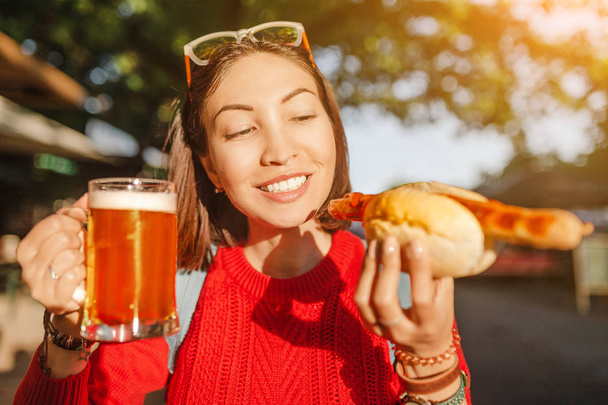 Young happy asian traveler woman drinks mug of beer with hotdog in Germany, beer and food festival concept - Photo, Image