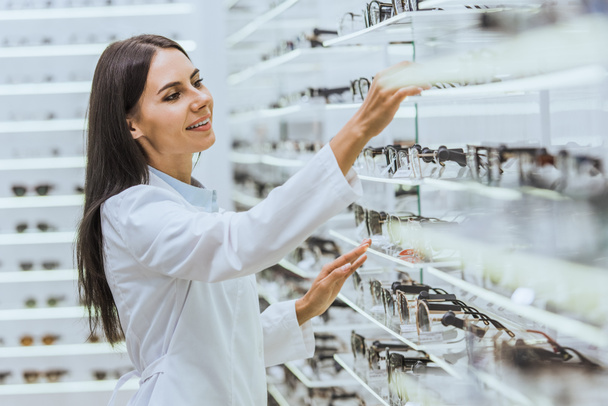 professional smiling optician taking glasses from shelves in ophthalmology - Photo, Image