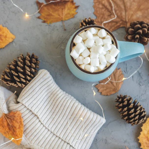 Blue mug with coffee, hot chocolate or cocoa with marshmallow on gray concrete background and lying dry leaves, cones, sweater, garland. Concept of warm cozy autumn or winter. - Valokuva, kuva