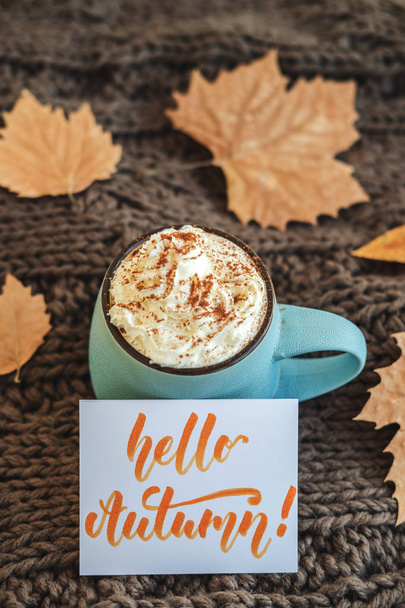 Mug of coffee, cocoa or hot chocolate with whipped cream and cinnamon on scarf with leaves, card Hello Autumn. Pumpkin latte - cozy drink for cold autumn or winter. - Фото, зображення