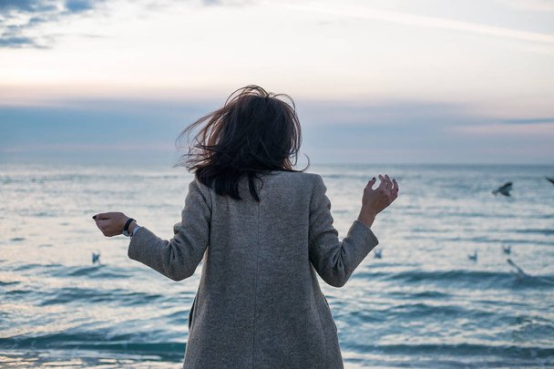 счастливая молодая женщина в сером пальто позирует на фоне моря, вид сзади
 - Фото, изображение