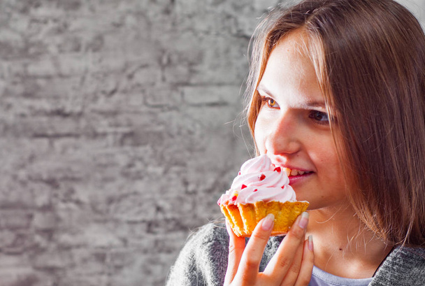 portrait of young teenager brunette girl with long hair eating cream cake on gray wall background - Photo, Image